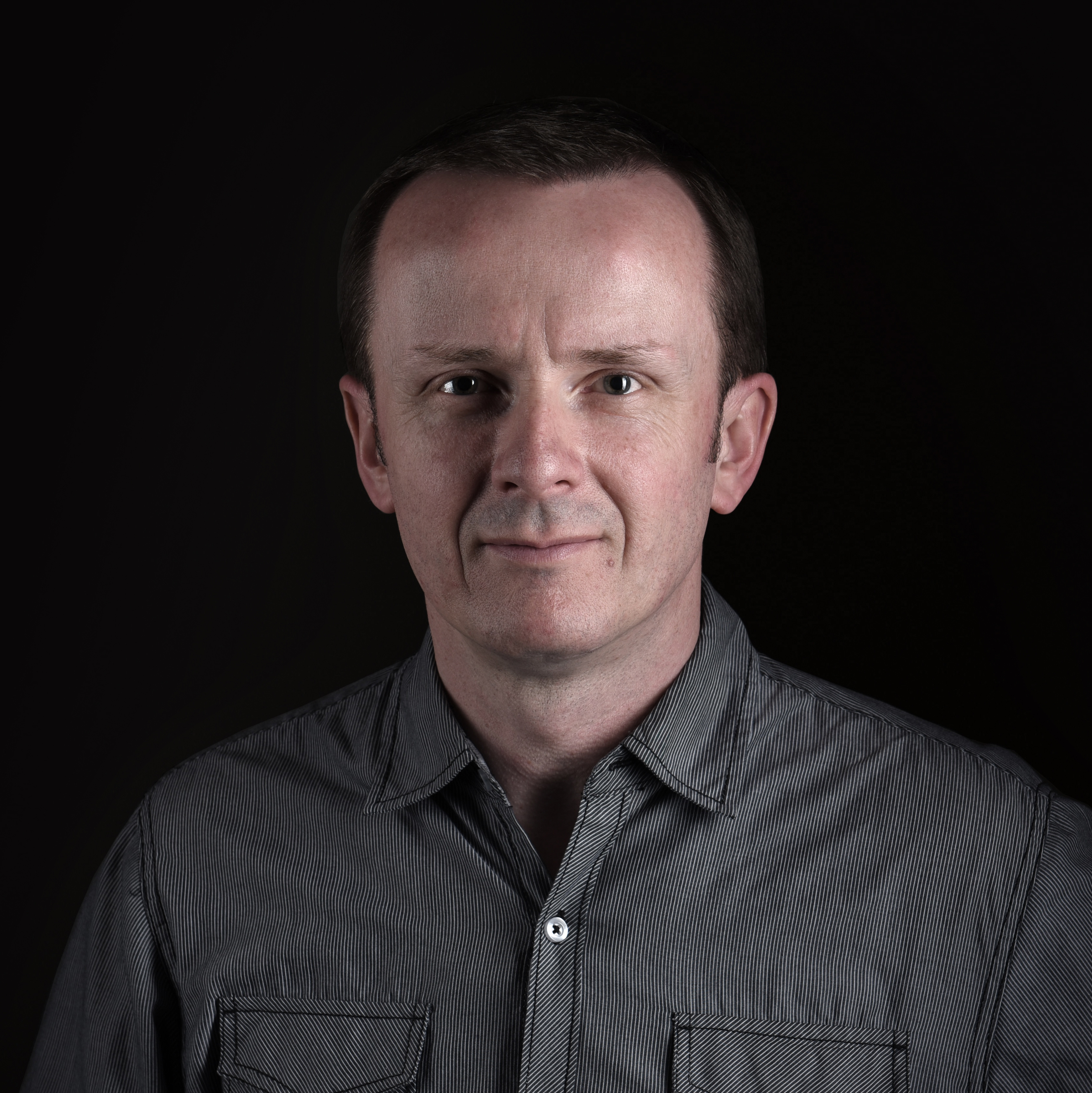 Man in patterned black and white shirt standing in front of a black background 