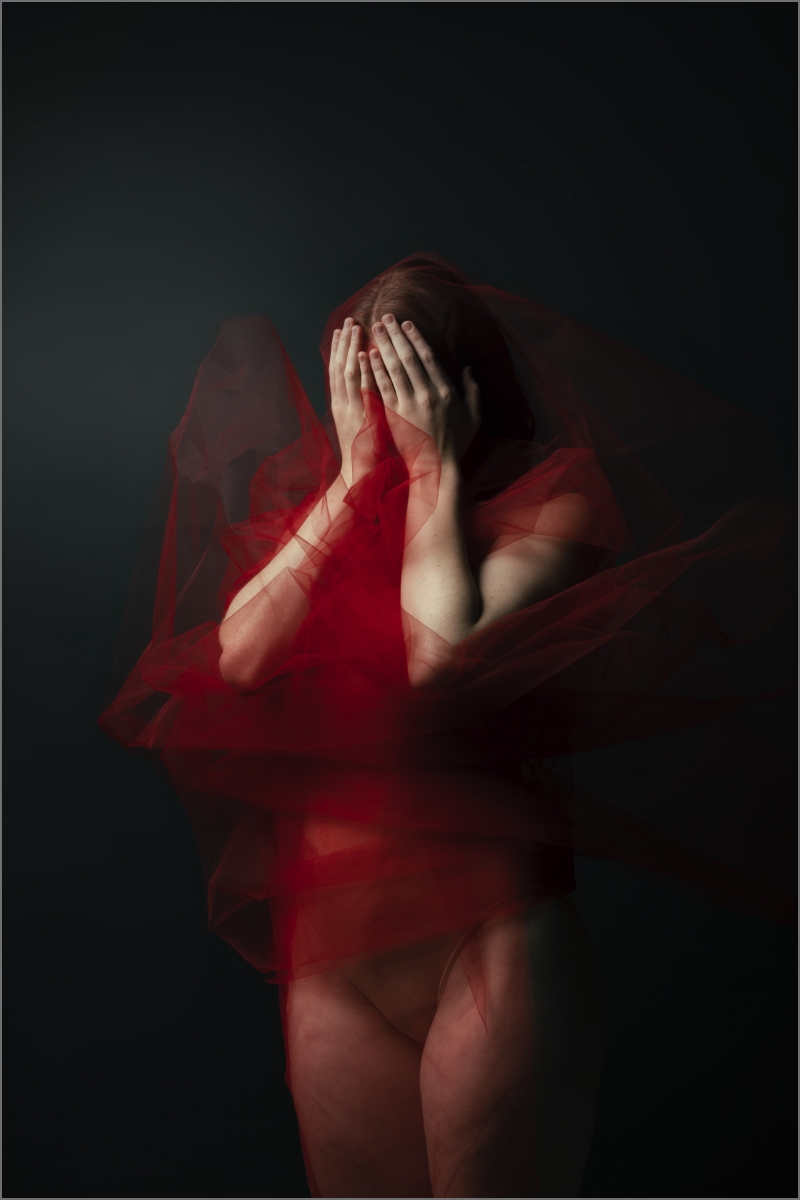 Person wrapped in red fabric with hands covering their faces against a dark background