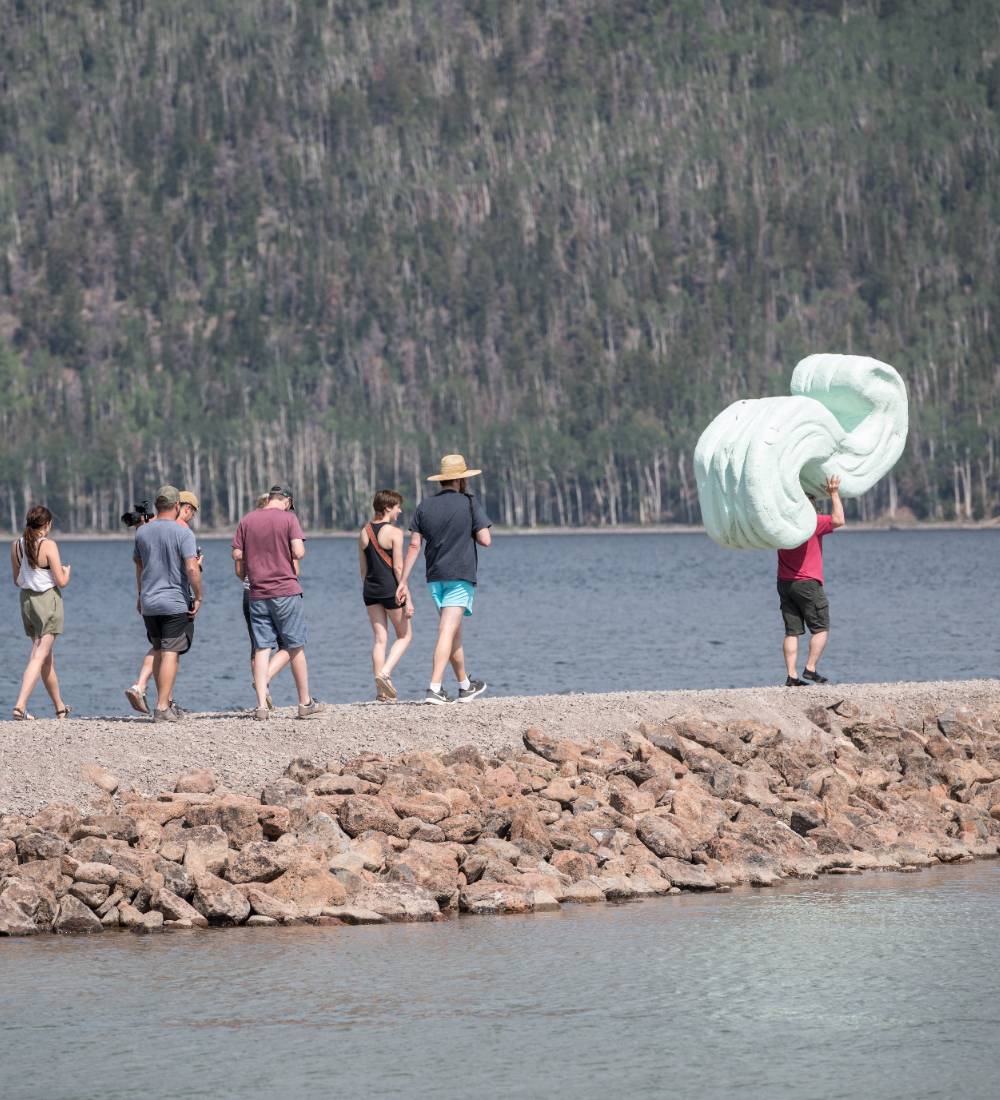 Six people walk along a stone path by a lake; one carries a large, light green sculpture.              