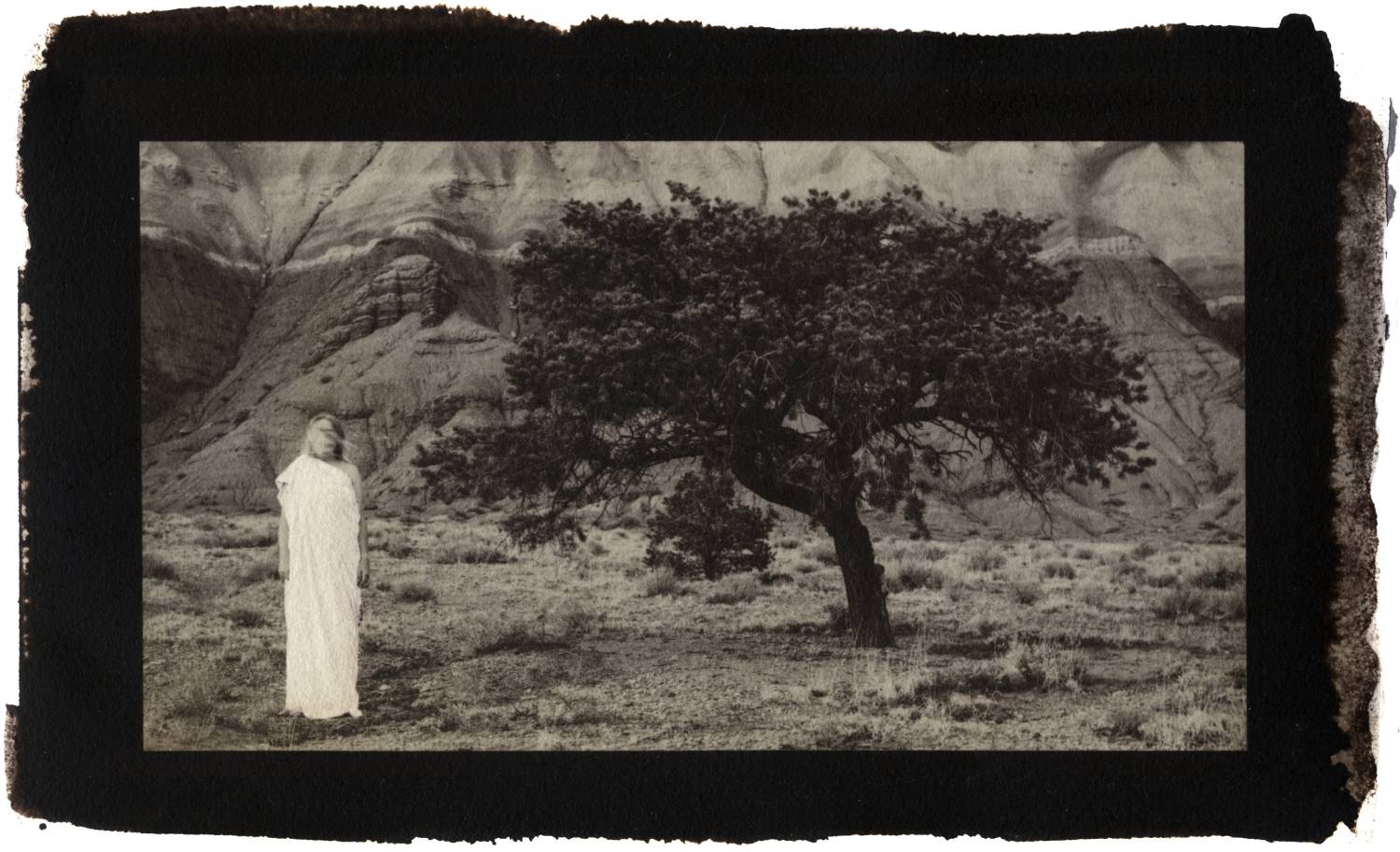 A sepia-tone photo of a person in a light robe next to a tree in a desert landscape with hills in the background.              