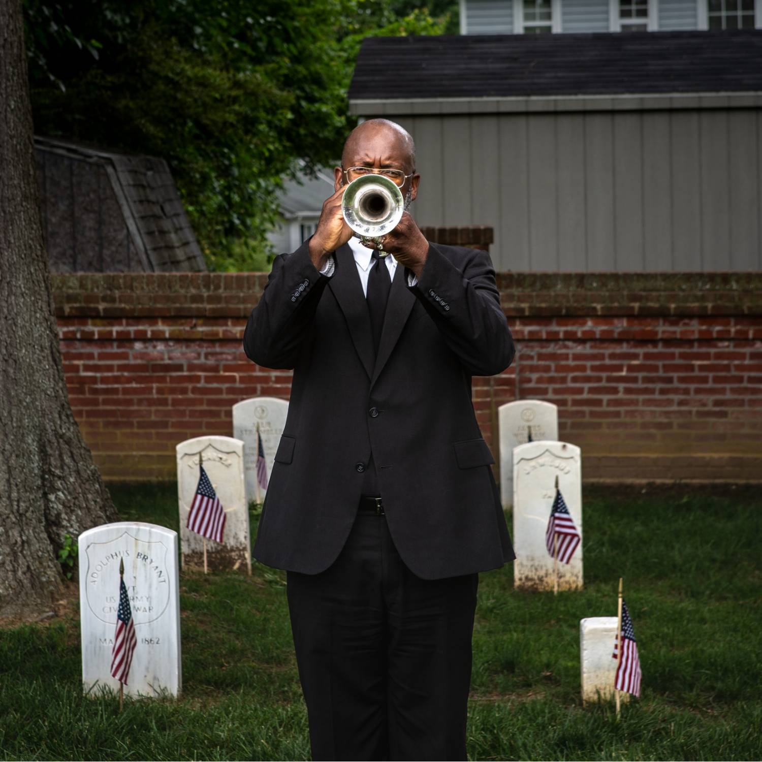 Man in a black suit playing a trumpet in a cemetery with military headstones and American flags.              