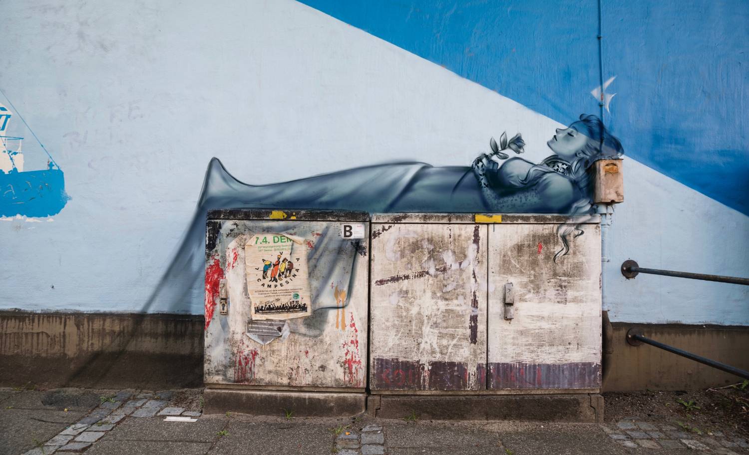 Street art of a reclining woman in gray hues on electrical boxes against a blue wall.              