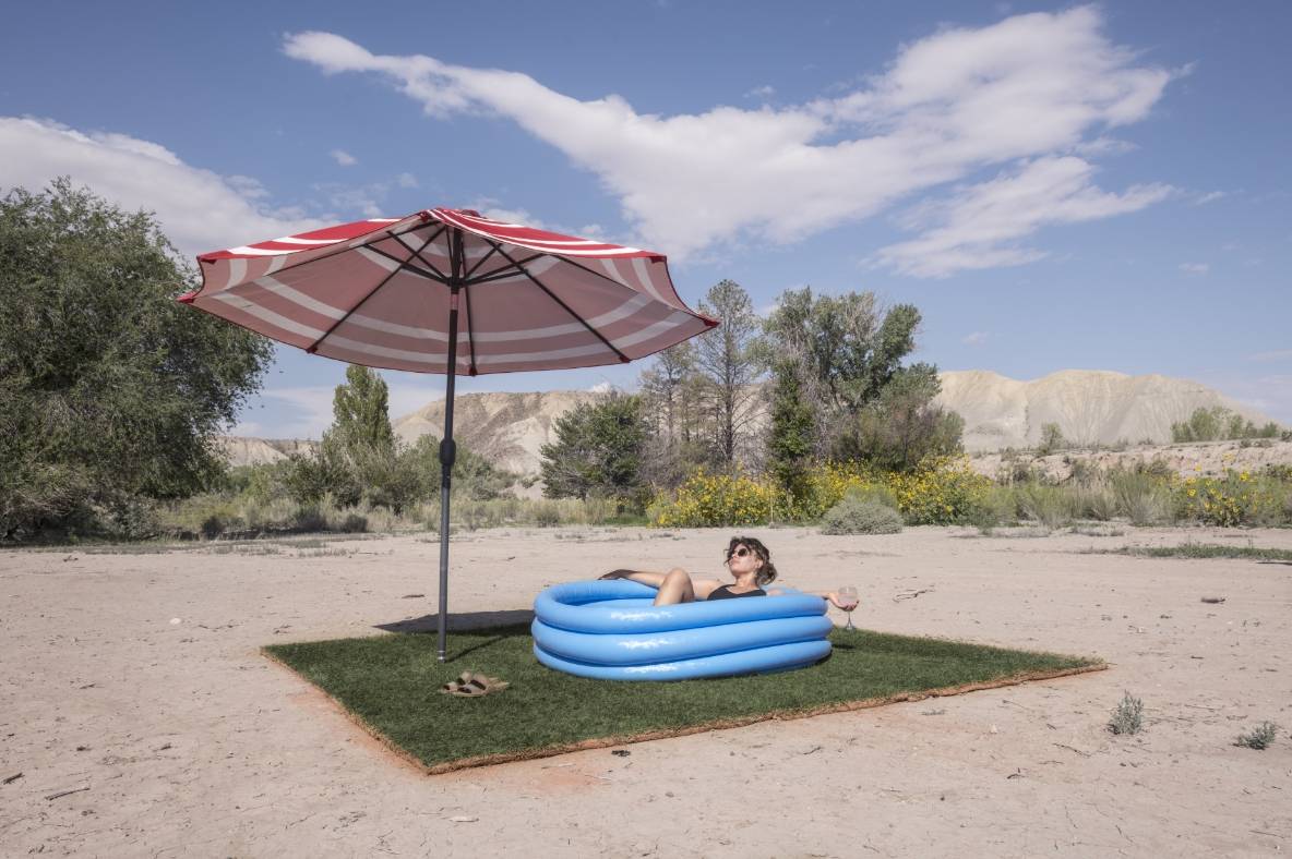 Person in a blue inflatable pool under a red and white umbrella on desert terrain.              