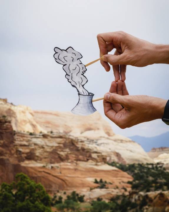 Hands holding a paper cut-out of a nuclear cooling tower with smoke in front of a desert landscape.              