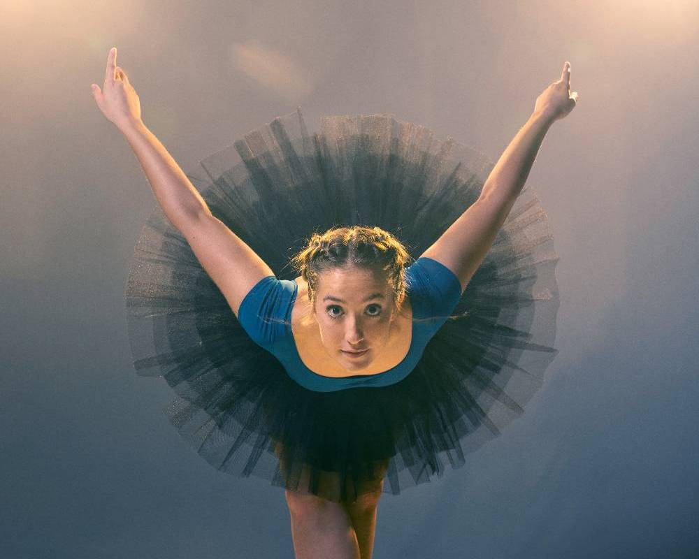 Ballet dancer in a black tutu with arms extended upwards in a V-shape, wearing a blue leotard.              