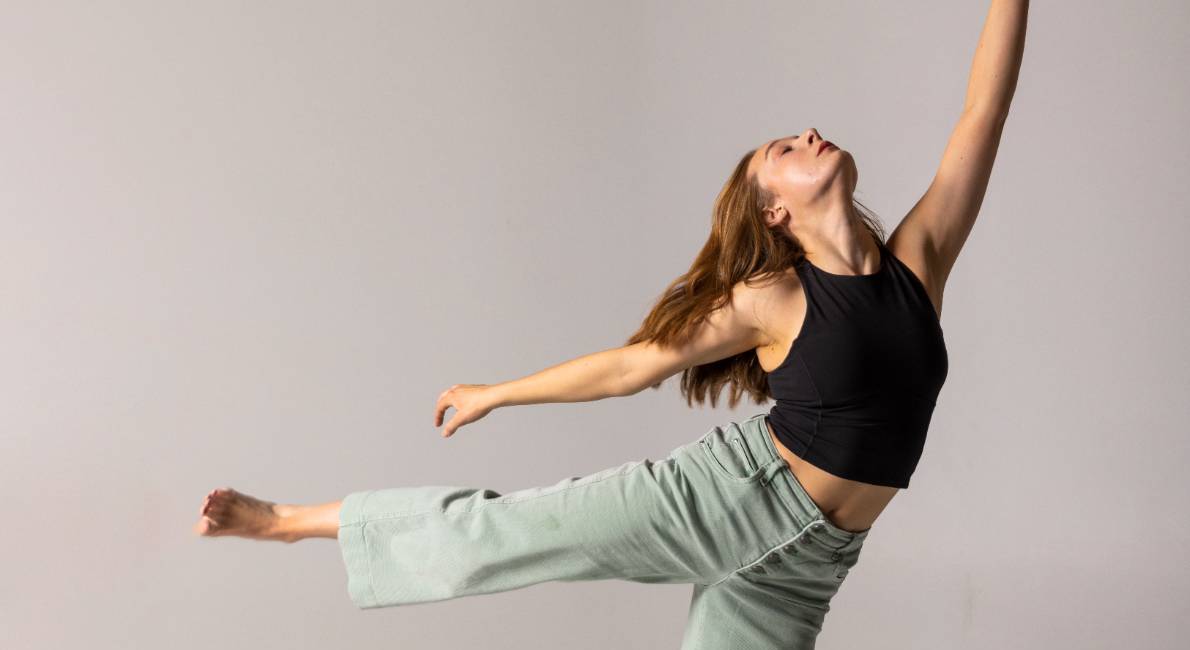 A dancer stretches her arm upward as she balances on one leg, wearing a black top and green pants.              