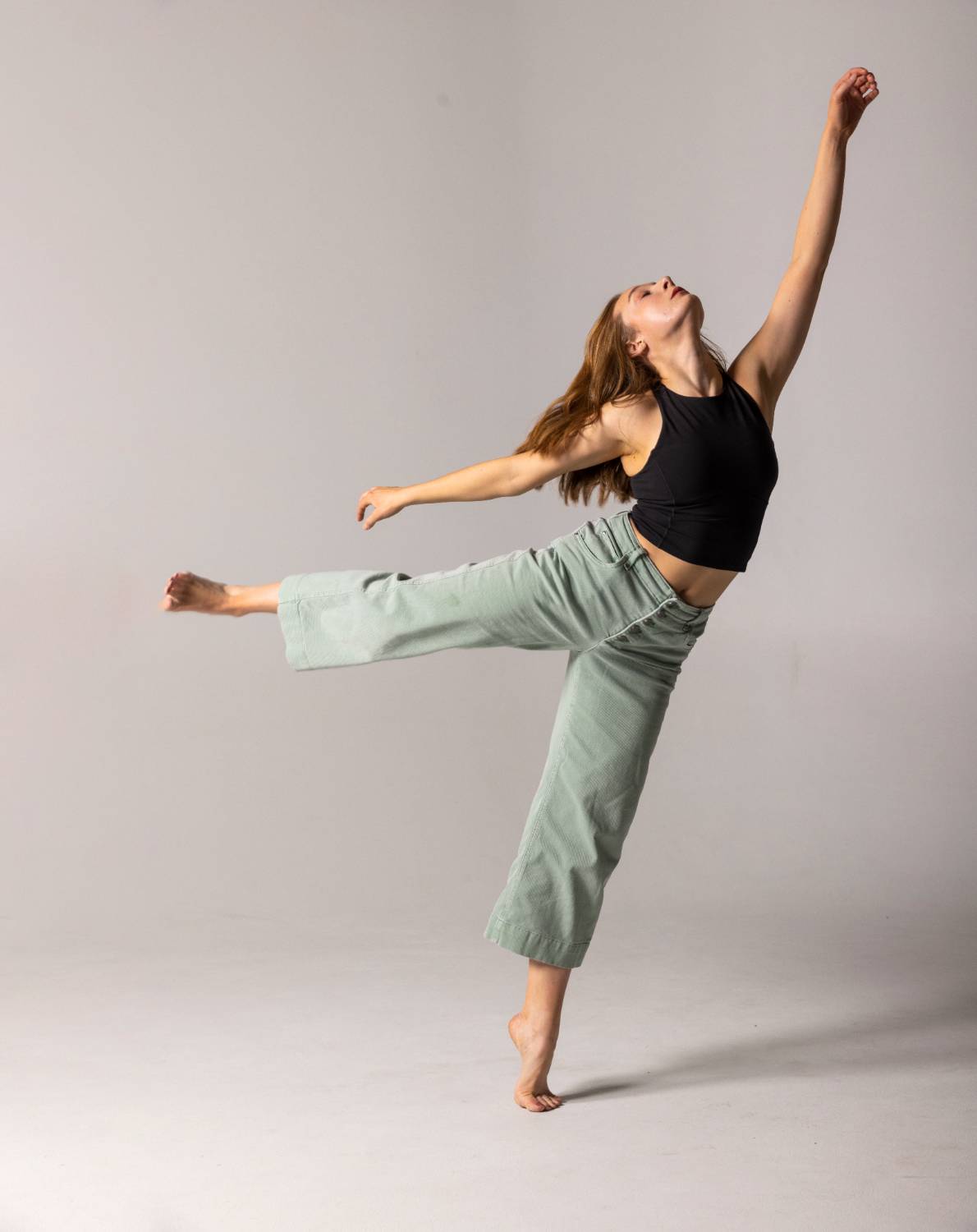 A dancer stretches her arm upward as she balances on one leg, wearing a black top and green pants.              