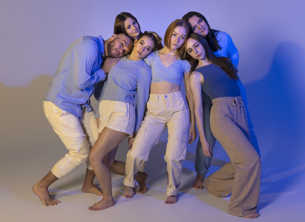 A group of six people in coordinated blue and beige outfits leaning together in a studio setting with a blue and purple gradient background.              