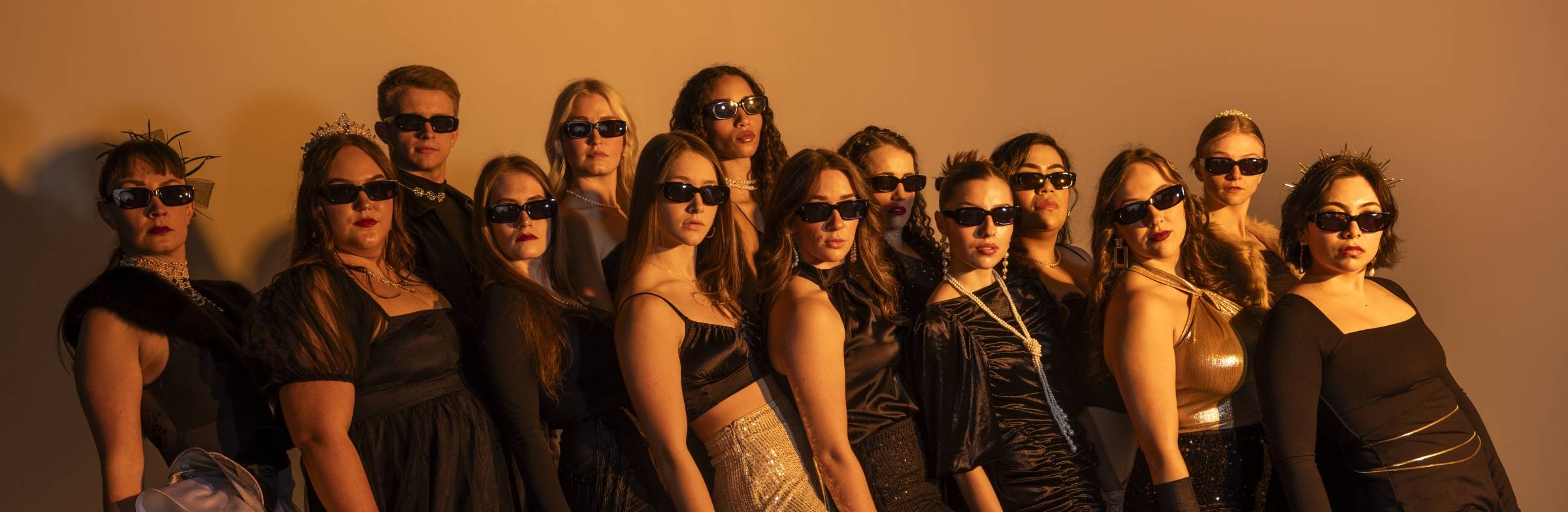 A group of fourteen people dressed in elegant black attire, all wearing dark sunglasses, posing together against a beige background.              