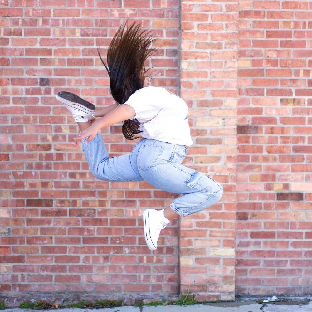 A person leaping in front of a red brick wall, wearing a white T-shirt, jeans, and white sneakers.              