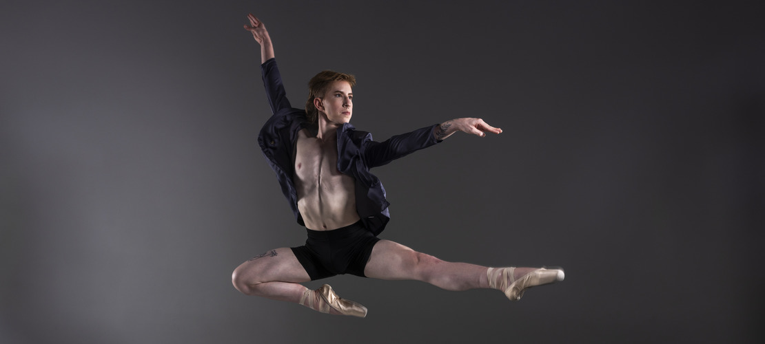 A ballet dancer performs a grand jeté mid-air against a gray background, wearing a dark jacket and black shorts.              