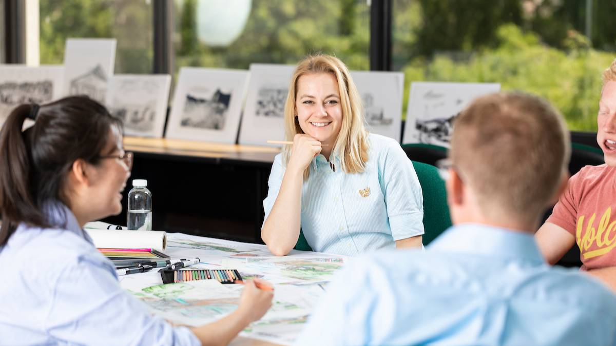 student smiling at camera with other group of students  looking at blueprints
