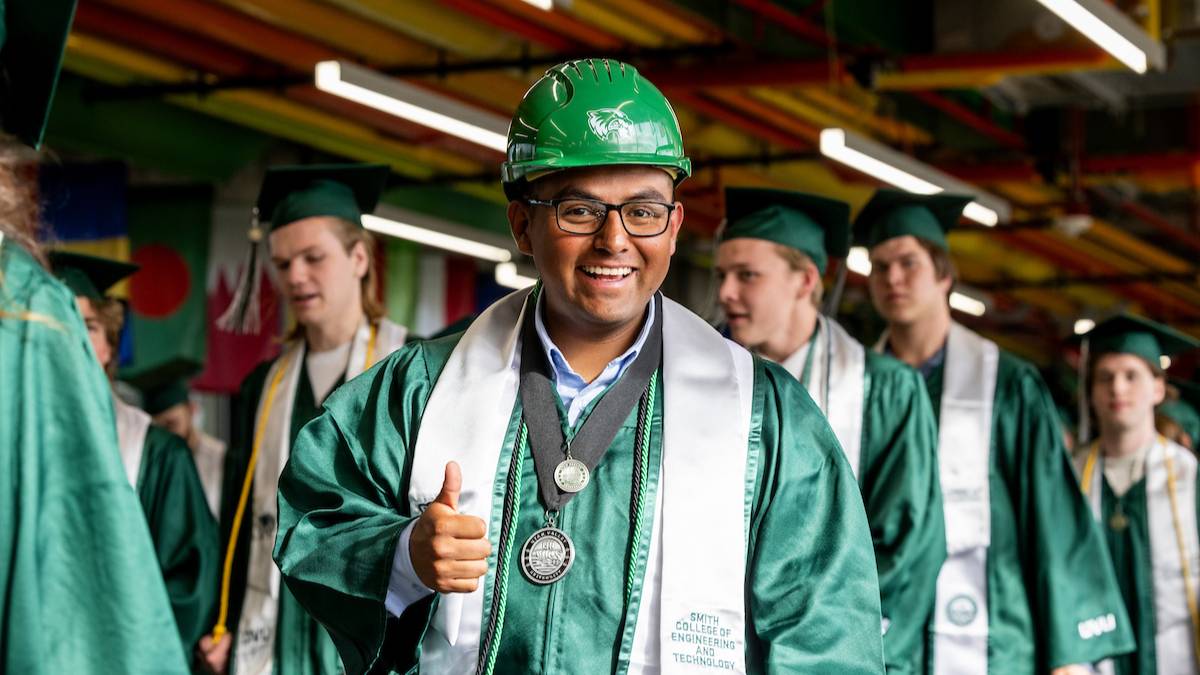 cuntruction student with uvu constuction hard hat and graduation gown smiling with a thumbs up