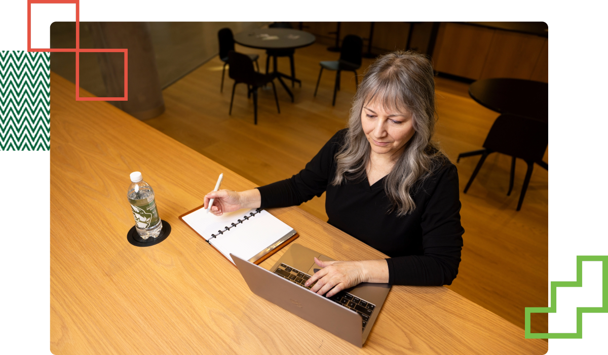 person writing notes while looking at their laptop in front of them