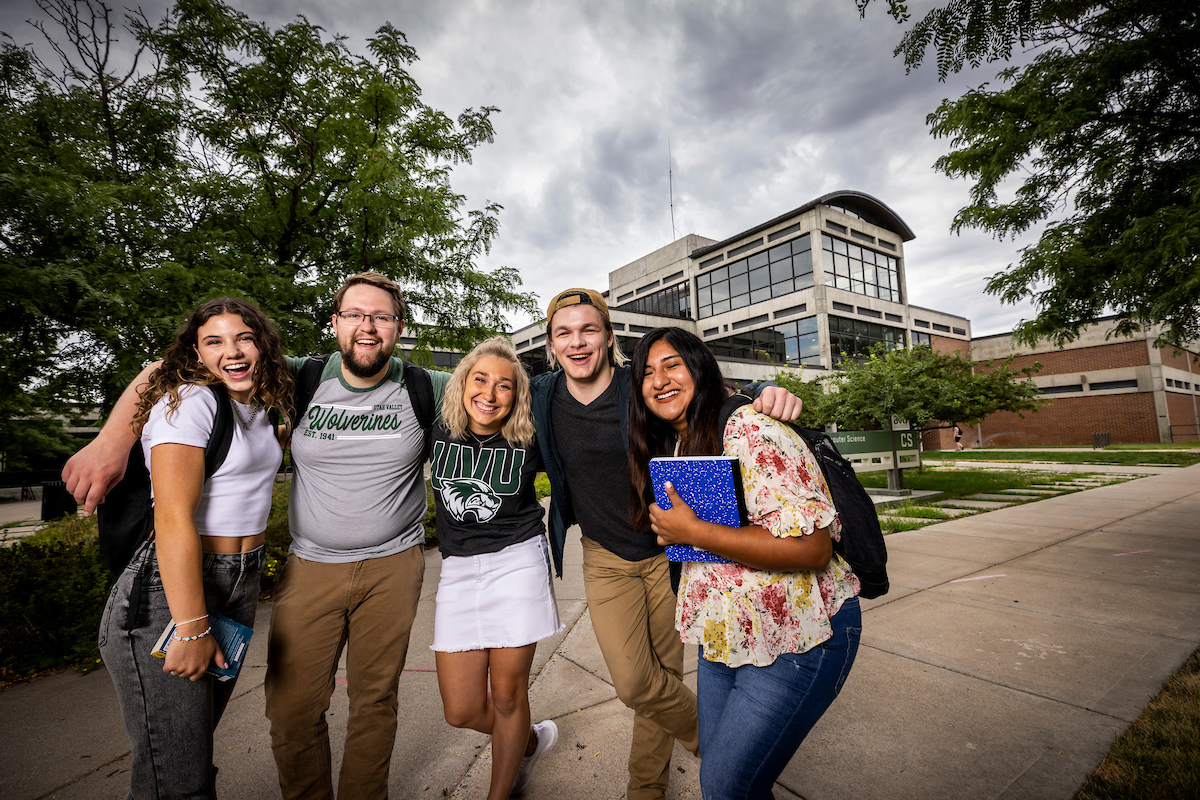Group of five smiling students standing with arms around each other in front of the Computer Science building on UVU campus.