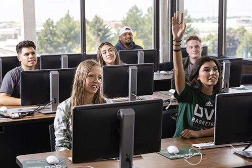 学生s behind individual computers sitting in a class as one student raises her hand
