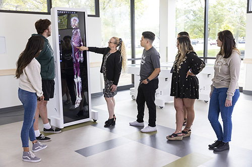 学生s being instructed by a teaching pointing at a six foot tall tablet